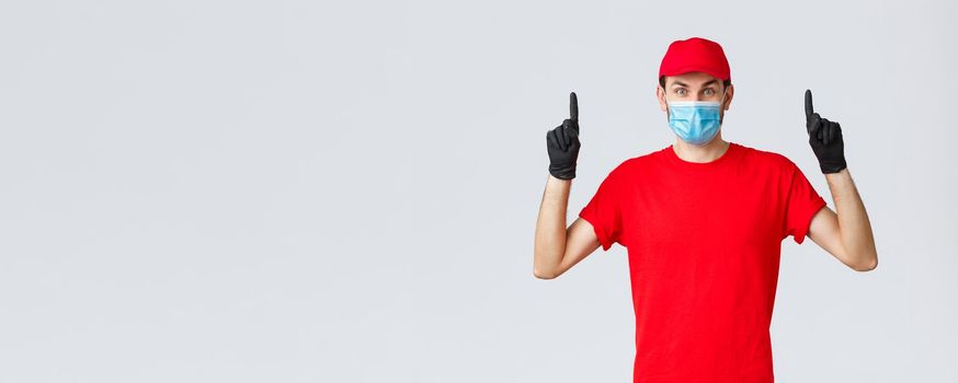 Covid-19, self-quarantine, online shopping and shipping concept. Surprised delivery guy in red uniform and cap, raising eyebrows amazed or excited, showing promo top, pointing up, wear medical mask.