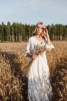 A blonde woman in a long white dress walks in a wheat field. The concept of a wedding and walking in nature.