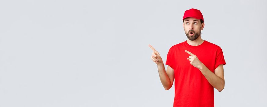 Online shopping, delivery during quarantine and takeaway concept. Impressed and wondered courier in red t-shirt with cap, employee uniform, open mouth shocked and staring left, pointing fingers.