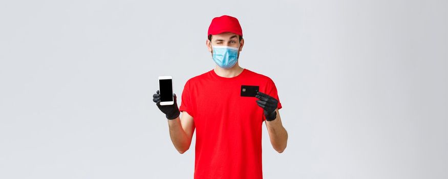 Contactless delivery, payment, online shopping during covid-19, self-quarantine. Handsome courier in red uniform, gloves and face mask, showing smartphone screen and credit card, promo of order app.