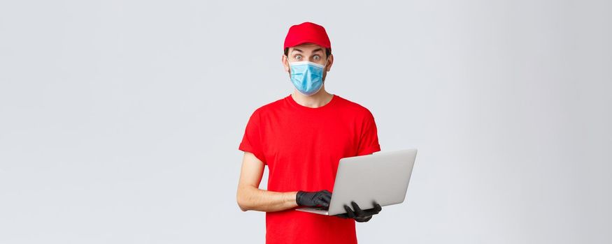 Customer support, covid-19 delivery packages, online orders processing concept. Surprised courier in red uniform, face mask and gloves, staring camera, holding laptop, read interesting news.