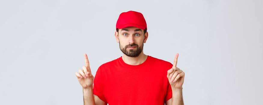 Online shopping, delivery during quarantine and takeaway concept. Indecisive and unsure courier in red uniform cap, t-shirt, smirk and pointing fingers up uncertain, dont know, cant decide.