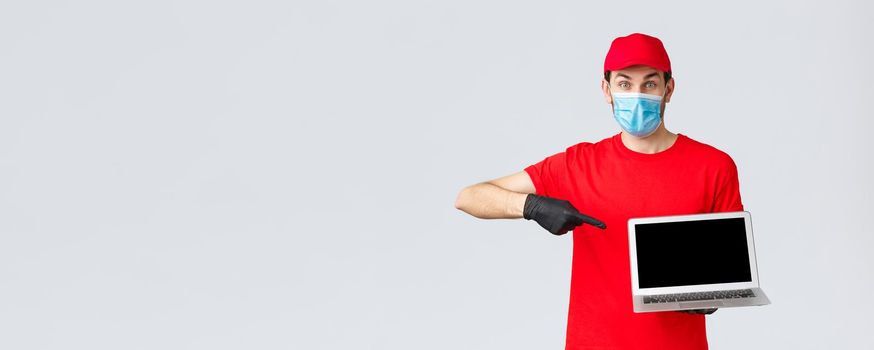 Customer support, covid-19 delivery packages, online orders processing concept. Enthusiastic courier in red uniform, gloves and face mask from coronavirus, pointing at laptop screen.