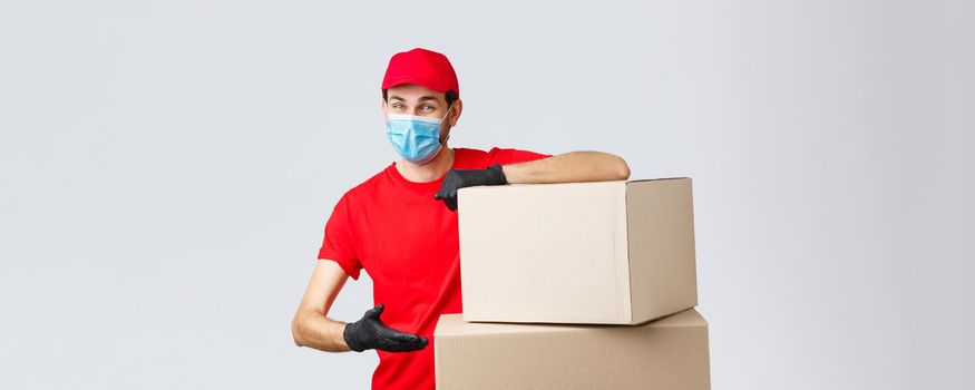 Packages and parcels delivery, covid-19 quarantine and transfer orders. Smiling courier in red uniform, gloves and medical face mask, introduce boxes to transfer your order, recommend service.