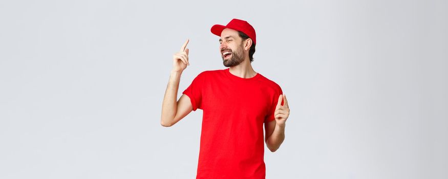 Good-looking carefree bearded man in red t-shirt and cap, uniform of employee, close eyes and dancing, pointing fingers up and singing. Delivery guy enjoying break at work, grey background.