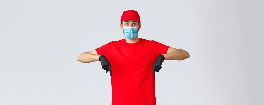 Covid-19, self-quarantine, online shopping and shipping concept. Surprised delivery guy in uniform pointing fingers down, showing promo. Courier in medical mask, gloves work with clients.