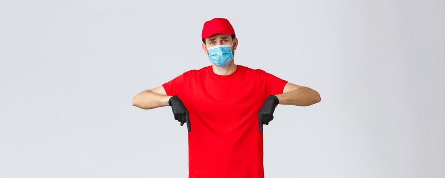 Covid-19, self-quarantine, online shopping and shipping concept. Upset and gloomy, sad delivery guy in red cap, t-shirt uniform, pointing fingers down disappointed. Courier showing bad news.