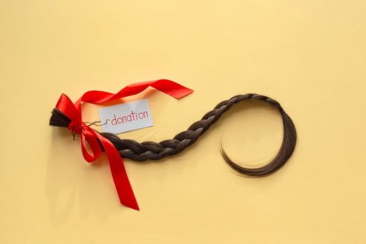 Long brown braid with tag label and red ribbon on yellow background, donation of haircut, natural human female hair, charity support and acts of kindess, top view