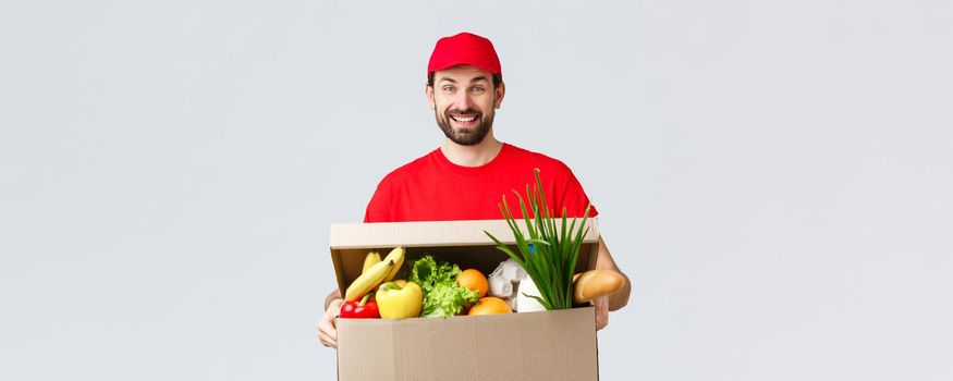 Groceries and packages delivery, covid-19, quarantine and shopping concept. Smiling delivery man in red uniform, bring client order, holding food box parcel, enjoying help people transfer goods.