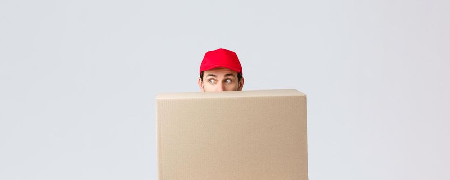 Packages and parcels delivery, covid-19 quarantine and transfer orders. Scared courier in red uniform cap, hiding behind customer order, looking left nervously, peeking at banner or advertisement.