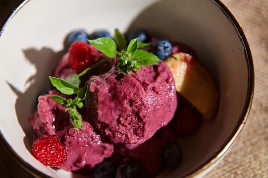 Homemade raw vegan ice cream, sorbet with blueberries, raspberries, ripe juicy peach slices and lemon basil leaves in a ceramic bowl. Healthy dessert. Rustic background. Food still life. Top view