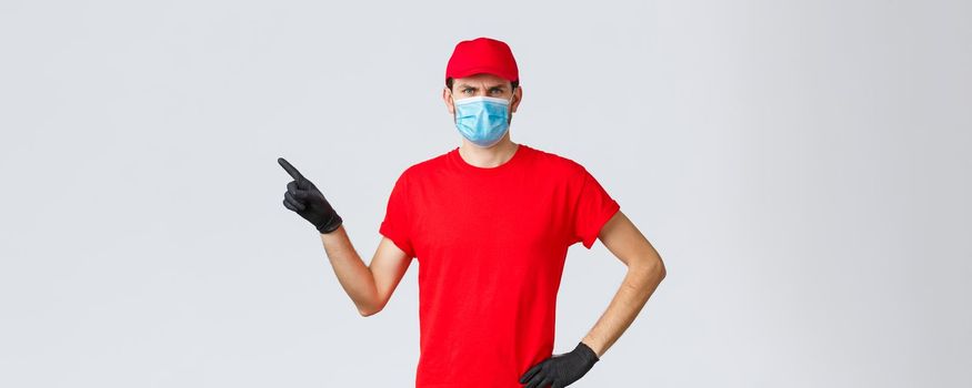 Covid-19, self-quarantine, online shopping and shipping concept. Displeased angry delivery guy scolding courier making mistake in transfer, frowning, pointing finger left, wear medical mask.