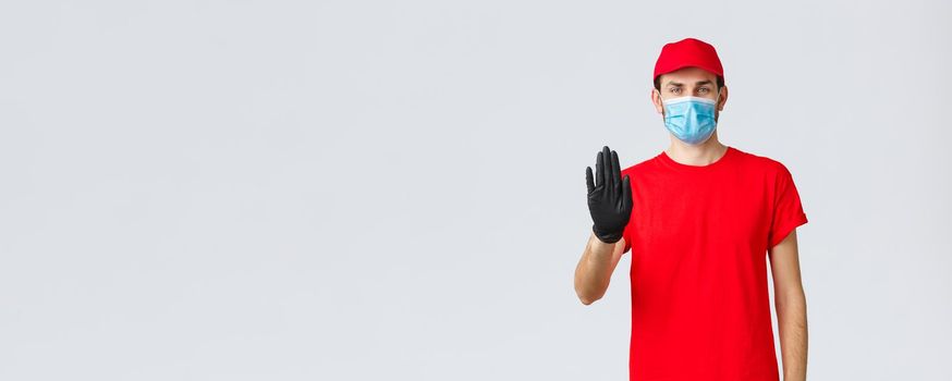 Covid-19, self-quarantine, online shopping concept. Confident delivery man in red uniform, gloves and face mask, raise hand in stop sign, prevent client step to storage without protective equipment.