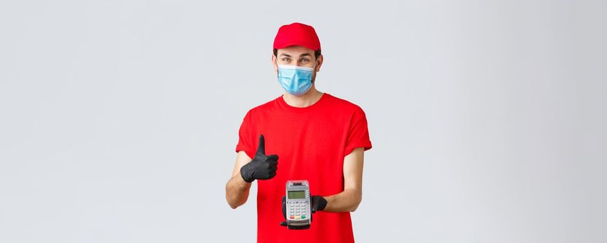 Contactless delivery, payment and online shopping during covid-19, self-quarantine. Friendly smiling courier in red uniform cap, t-shrit, medical mask and gloves, advice pay order with POS terminal.