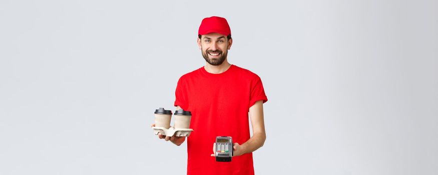 Food delivery, quarantine, stay home and order online concept. Friendly bearded courier in red uniform handing coffee delivery and POS terminal for client to pay contactless, grey background.