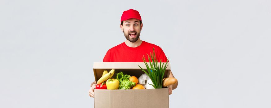 Groceries and packages delivery, covid-19, quarantine and shopping concept. Smiling handsome bearded courier in red uniform, bring food package, grocery order to client in box, look amused.