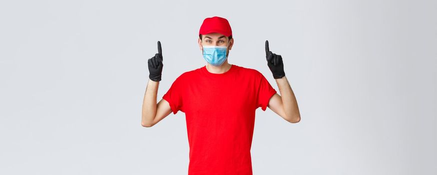 Covid-19, self-quarantine, online shopping and shipping concept. Amused happy delivery guy advertise courier company service during coronavirus lockdown, pointing fingers up, wear medical mask.