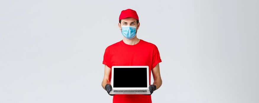 Customer support, covid-19 delivery packages, online orders processing concept. Smiling courier in red uniform, face mask and gloves, showing laptop screen webpage of company.