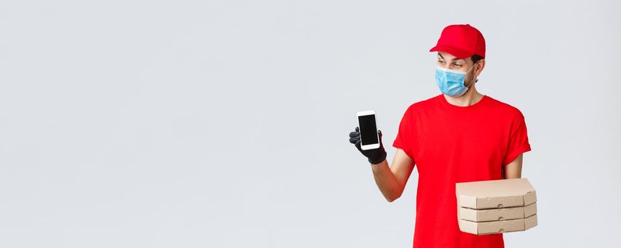 Food delivery, application, online grocery, contactless shopping and covid-19 concept. Surprised smiling courier in red uniform, holding pizza boxes and smartphone, showing app for bonuses or deliver.