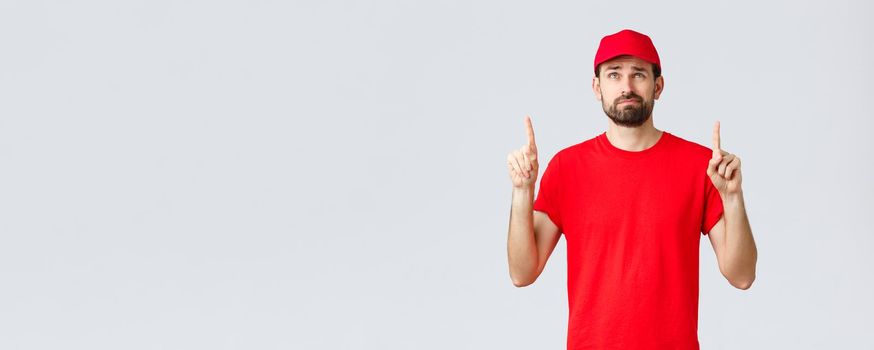 Online shopping, delivery during quarantine and takeaway concept. Displeased, reluctant young courier, employee in red uniform cap and t-shirt, smirk uncertain, reading bad news upwards, point up.