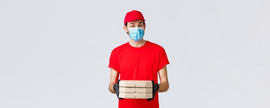 Food delivery, application, online grocery, contactless shopping and covid-19 concept. Pleasant courier in red uniform bring pizza in boxes, wear face mask and rubber gloves, give clint order.