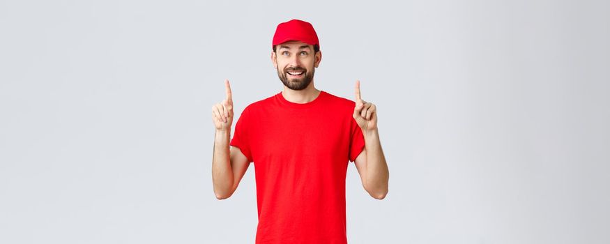 Online shopping, delivery during quarantine and takeaway concept. Smiling cheerful courier in red t-shirt, cap pointing fingers up. Employee look intrigued at banner with special discount.