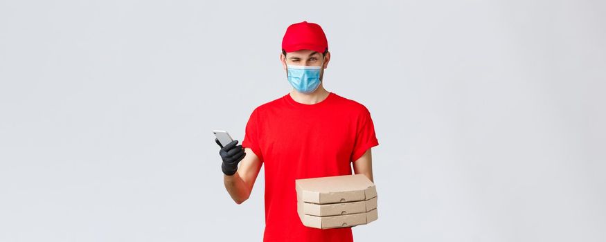 Food delivery, application, online contactless shopping and covid-19 concept. Courier in red uniform, face mask and gloves, winking to client, inform bonuses, special discounts on pizza, hold phone.