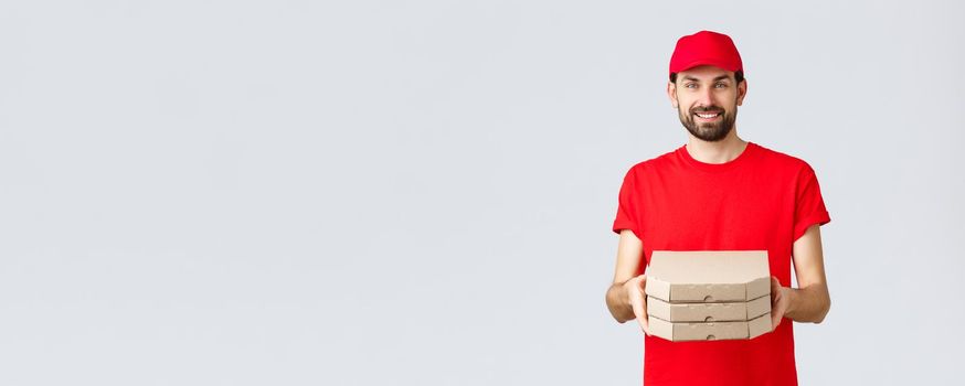 Food delivery, quarantine, stay home and order online concept. Smiling nice bearded courier in red uniform cap and t-shirt, handing clients boxes with pizza, deliver order, grey background.