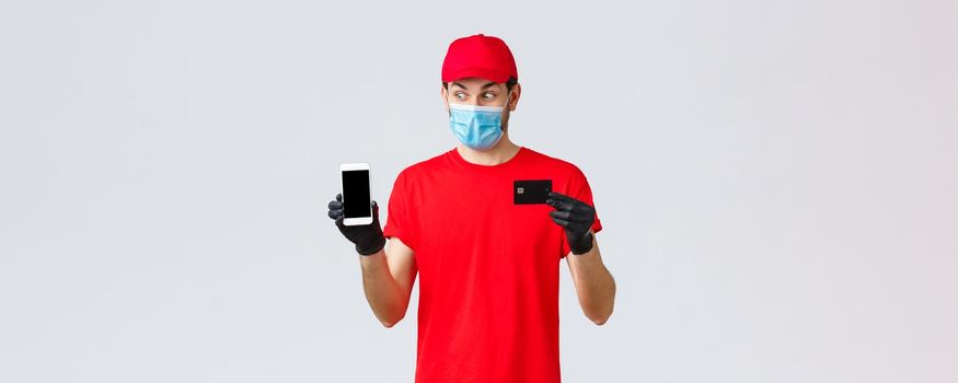 Contactless delivery, payment and online shopping during covid-19, self-quarantine. Excited courier in red uniform, face mask and gloves, look smartphone display, show credit card, paying order.