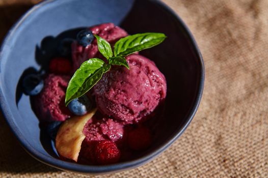 Top view. Food still life. Delicious healthy raw vegan berry ice cream sorbet, with ripe juicy fruits, blueberries and lemon basil leaves in a dark blue ceramic bowl on a linen tablecloth. Copy space