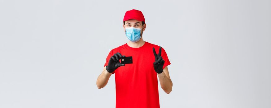 Contactless delivery, payment and online shopping during covid-19, self-quarantine. Friendly courier in red uniform, gloves and face mask provide safe paying option, show credit card and peace sign.