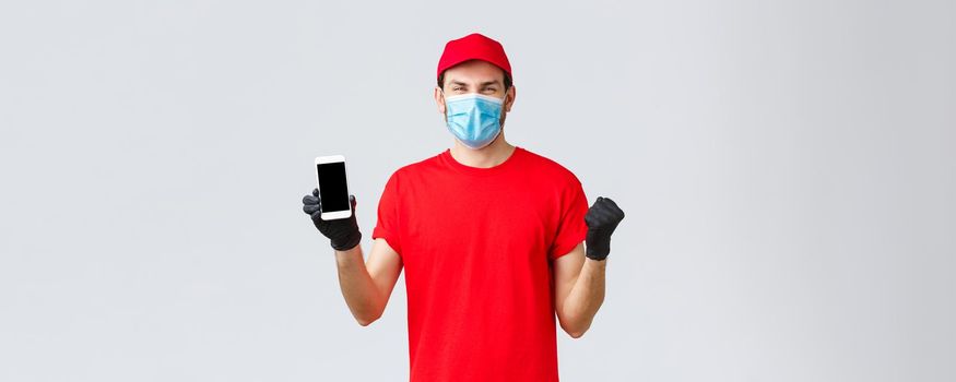 Contactless delivery, payment and online shopping during covid-19, self-quarantine. Rejoicing cheerful courier in red uniform cap, t-shirt celebrating amazing promo, show smartphone screen, wear mask.
