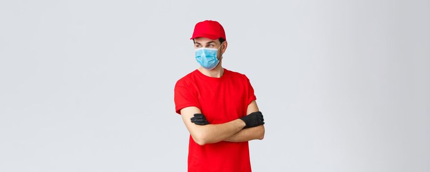 Covid-19, self-quarantine, online shopping and shipping concept. Confident courier in red uniform, protective gloves and face mask, standing crossed arms, look away, ready take your orders.