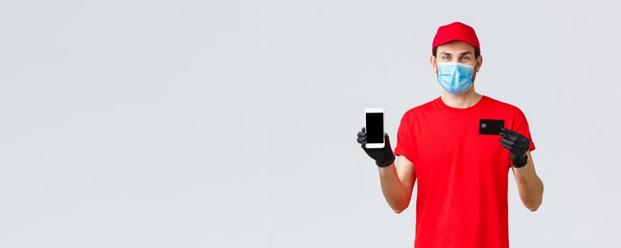Contactless delivery, payment and online shopping during covid-19, self-quarantine. Smiling courier in red uniform, gloves and face mask showing smartphone screen and credit card, app for orders.
