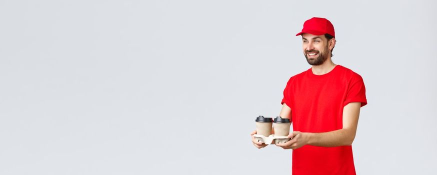 Food delivery, quarantine, stay home and order online concept. Friendly courier in red uniform cap and t-shirt, handing coffee to clients, bring bevereges via drive-through window, grey background.