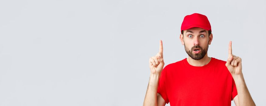 Online shopping, delivery during quarantine and takeaway concept. Surprised and impressed delivery guy, courier in red t-shirt and cap, pointing fingers up, gasping amazed, read interesting info.