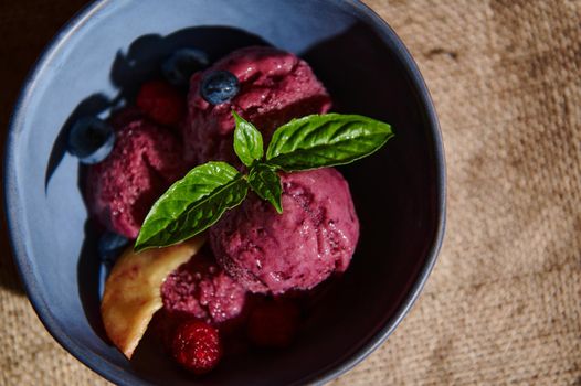 Flat lay. Food still life. Delicious healthy raw vegan berry ice cream sorbet, with ripe juicy fruits, blueberries and lemon basil leaves in a dark blue ceramic bowl on a linen tablecloth. Copy space