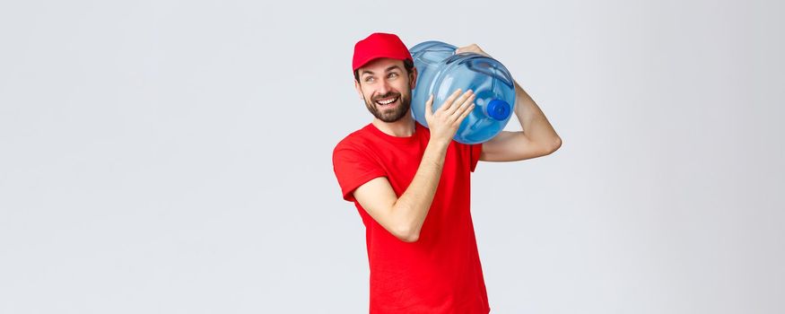 Takeaway, food and groceries delivery, covid-19 contactless orders concept. Cheerful smiling bearded courier in red uniform, cap, bring bottled water to office or house, look away joyful.