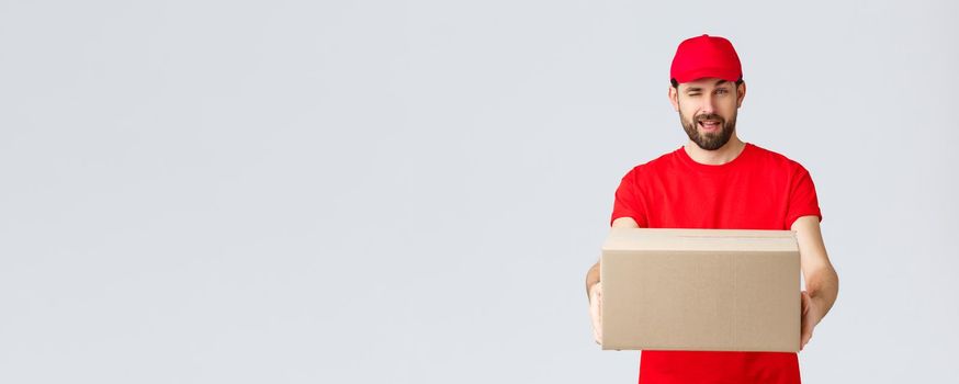 Order delivery, online shopping and package shipping concept. Cheeky handsome bearded courier in red uniform, handing box package to client. Employee wink to you and give order parcel.