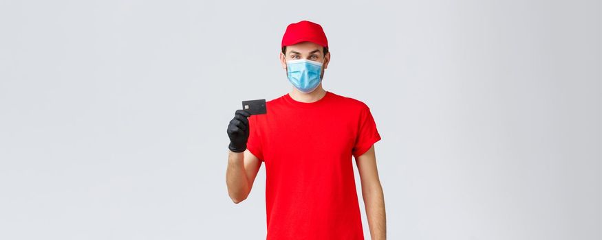 Contactless delivery, payment and online shopping during covid-19, self-quarantine. Young courier in red uniform cap, face mask and gloves, showing credit card, easy paying and ordering.
