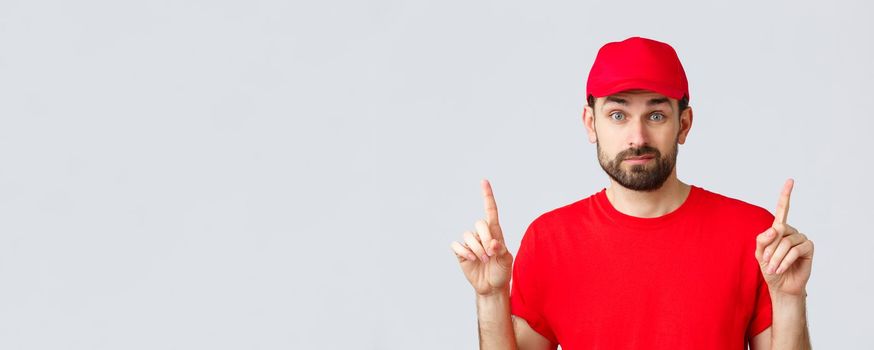 Online shopping, delivery during quarantine and takeaway concept. Indecisive and unsure courier in red uniform cap, t-shirt, smirk and pointing fingers up uncertain, dont know, cant decide.