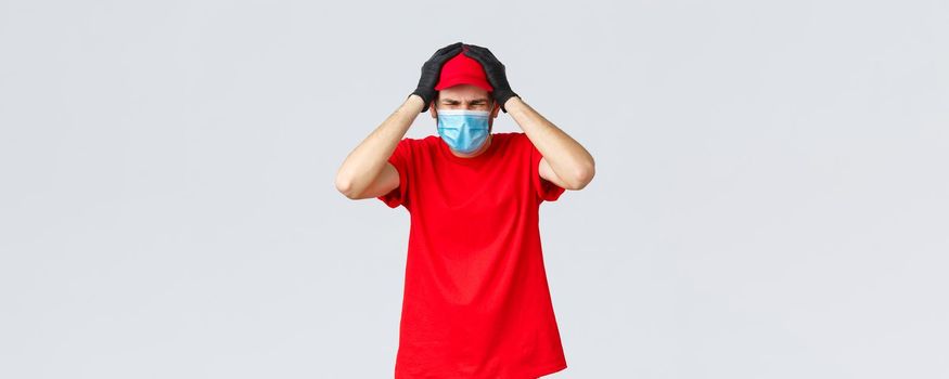 Covid-19, self-quarantine, online shopping and shipping concept. Troubled and embarrassed courier man in medical mask, gloves, hold hands on head, grimacing, making mistake with parcel delivery.