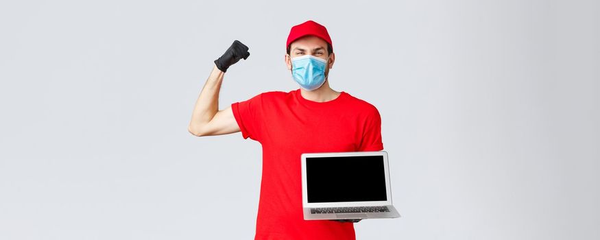 Customer support, covid-19 delivery packages, online orders processing concept. Rejoicing happy courier lift hand in yes, celebration, showing laptop screen, wear face mask and gloves.