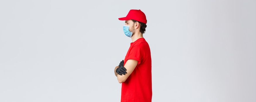 Covid-19, self-quarantine, online shopping and shipping concept. Profile f determined courier in red t-shirt and cap, uniform of carrier, cross hands chest, working shift, give-out clients orders.