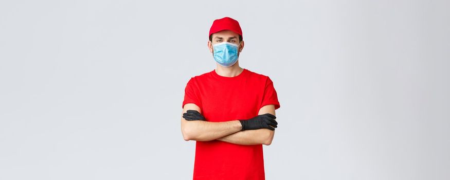 Confident young delivery guy in red t-shirt and cap, wearing medical mask and protective gloves while delivering orders to customers staying home safe during coronavirus self-quarantine.