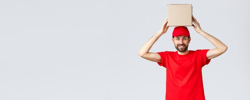 Order delivery, online shopping and package shipping concept. Funny and cute bearded courier in red uniform cap and t-shirt, holding box on head. Employee with package smiling.