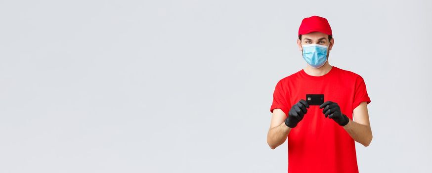 Contactless delivery, payment and online shopping during covid-19, self-quarantine. Cheerful courier in red uniform cap and t-shirt, wear medical face mask, gloves showing credit card, order food.