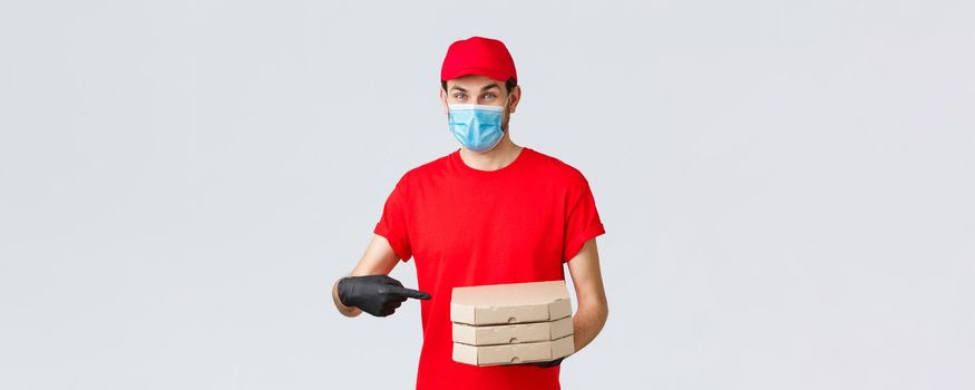 Food delivery, application, online grocery, contactless shopping and covid-19 concept. Cheerful courier in red uniform, face mask and gloves pointing finger at pizza boxes, deliver to client house.