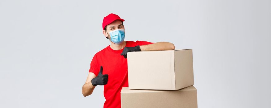 Packages and parcels delivery, covid-19 quarantine and transfer orders. Confident courier in red uniform, gloves and medical mask, encourage call service, show thumb-up lean on boxes.