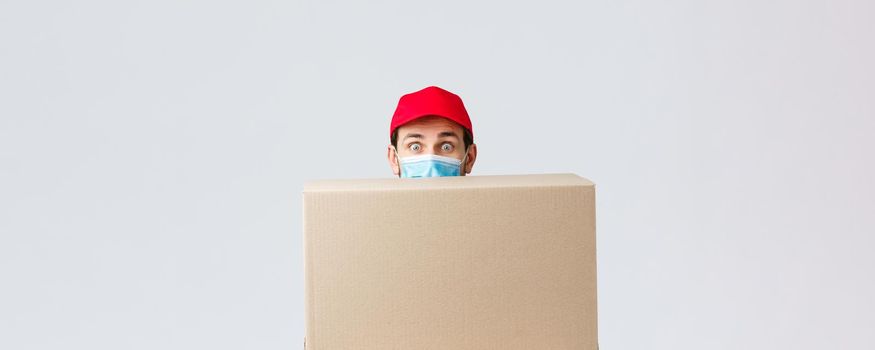 Packages and parcels delivery, covid-19 quarantine and transfer orders. Clumsy cute courier first day at job, hiding behind large box, wear face mask and red uniform cap, grey background.